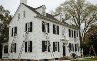 Painters on ladders repaint a large white house with black shutters.